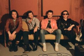 Portrait of Arctic Monkeys sitting on low sofa against a brown wall.