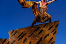Stephenson Ardern-Sodje in costume as the Lion King, standing atop an orange stage set cliff with arms outstretched.