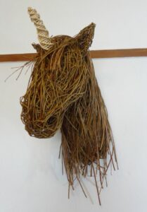 Picture shows unicorn head made of dark willow with paler horn, mounted on white wall.