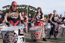 Photo shows three drummers dressed in red and black at Victorious Festival. Katie is in the centre of the picture, with a drum from waist to ankles, and a big smile.