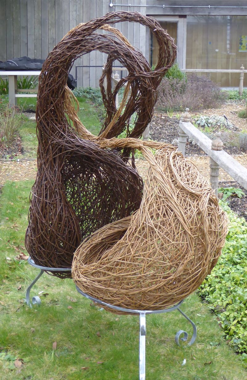 Picture shows organic willow sculpture, similar to yin yang, made of darker and lighter woven willow, mounted on base in garden.