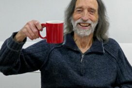 Bearded man with dark hair raising a mug a smiling at camera.