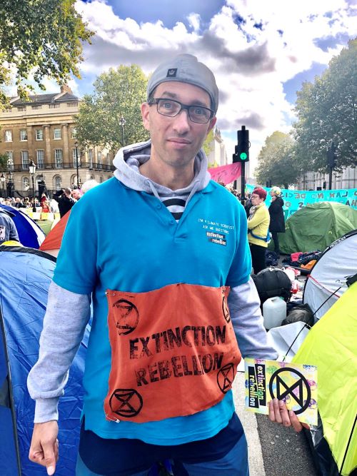 Bors Hulesch in Extinction Rebellion top at a rally, holding XR leaflets and looking into camera.